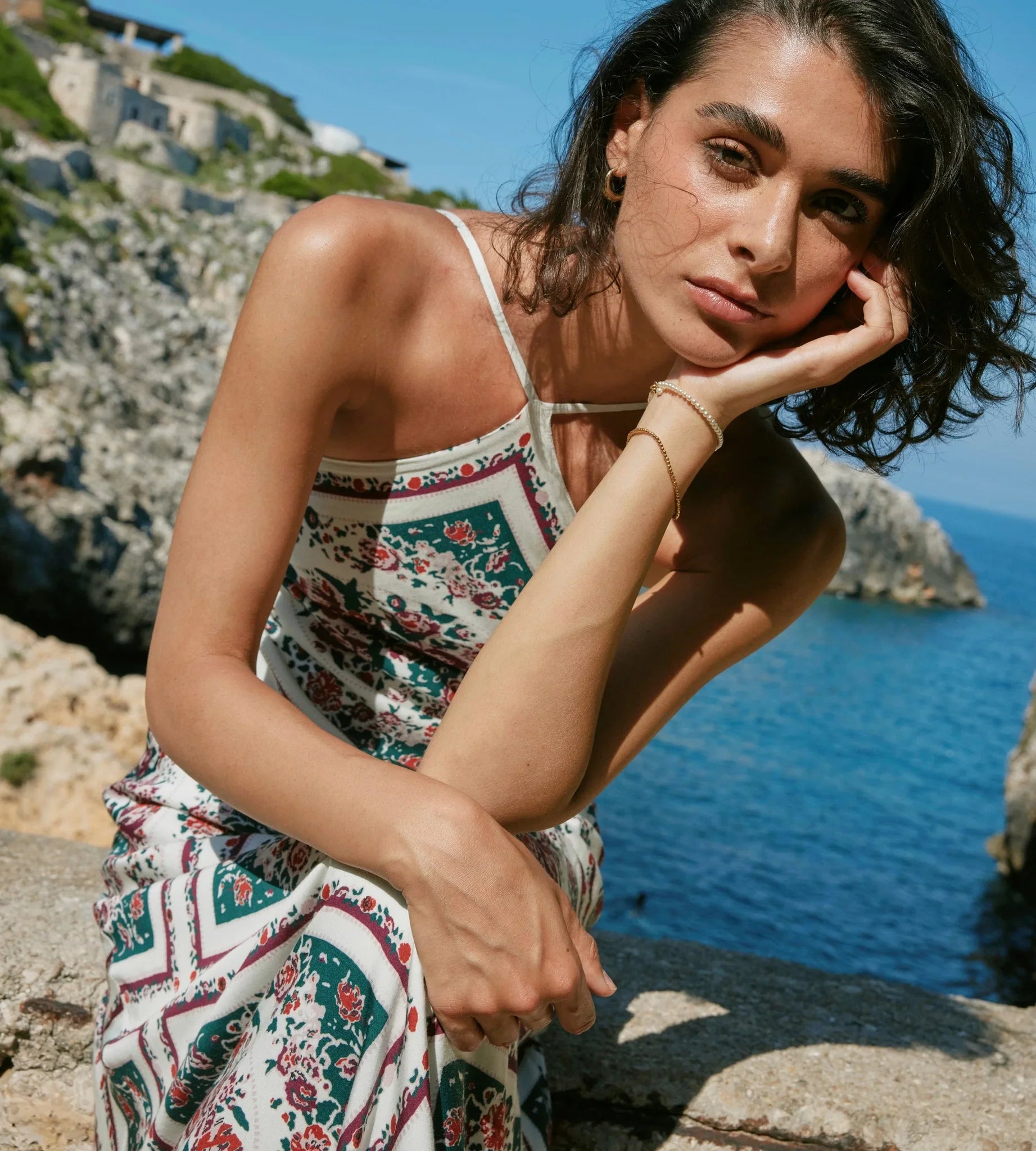 A woman wearing a white spaghetti-strap dress with a red, green, and blue floral pattern sits on a rocky coastal cliff with the blue sea and sky in the background.