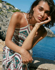 A woman wearing a white spaghetti-strap dress with a red, green, and blue floral pattern sits on a rocky coastal cliff with the blue sea and sky in the background.