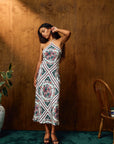 A woman models a sleeveless halter midi dress with a colorful geometric and floral mandala pattern, standing in front of a rustic wooden wall.