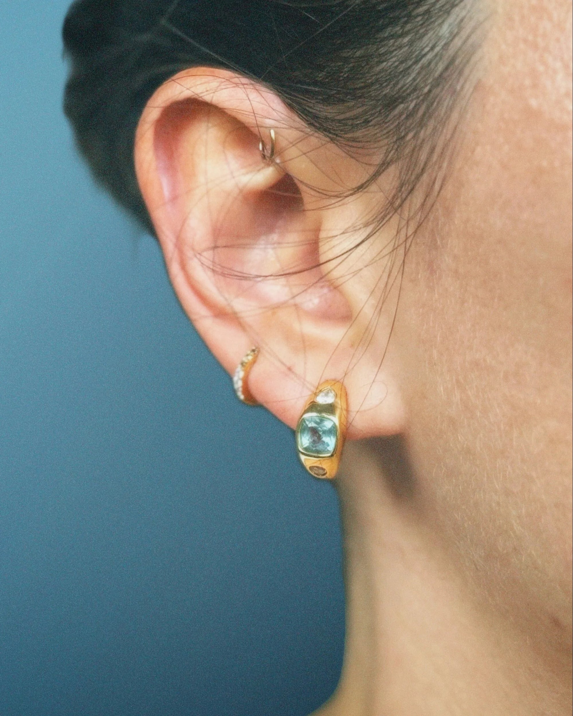 Close-up of an ear wearing gold hoop earrings with blue gemstones against a blue background