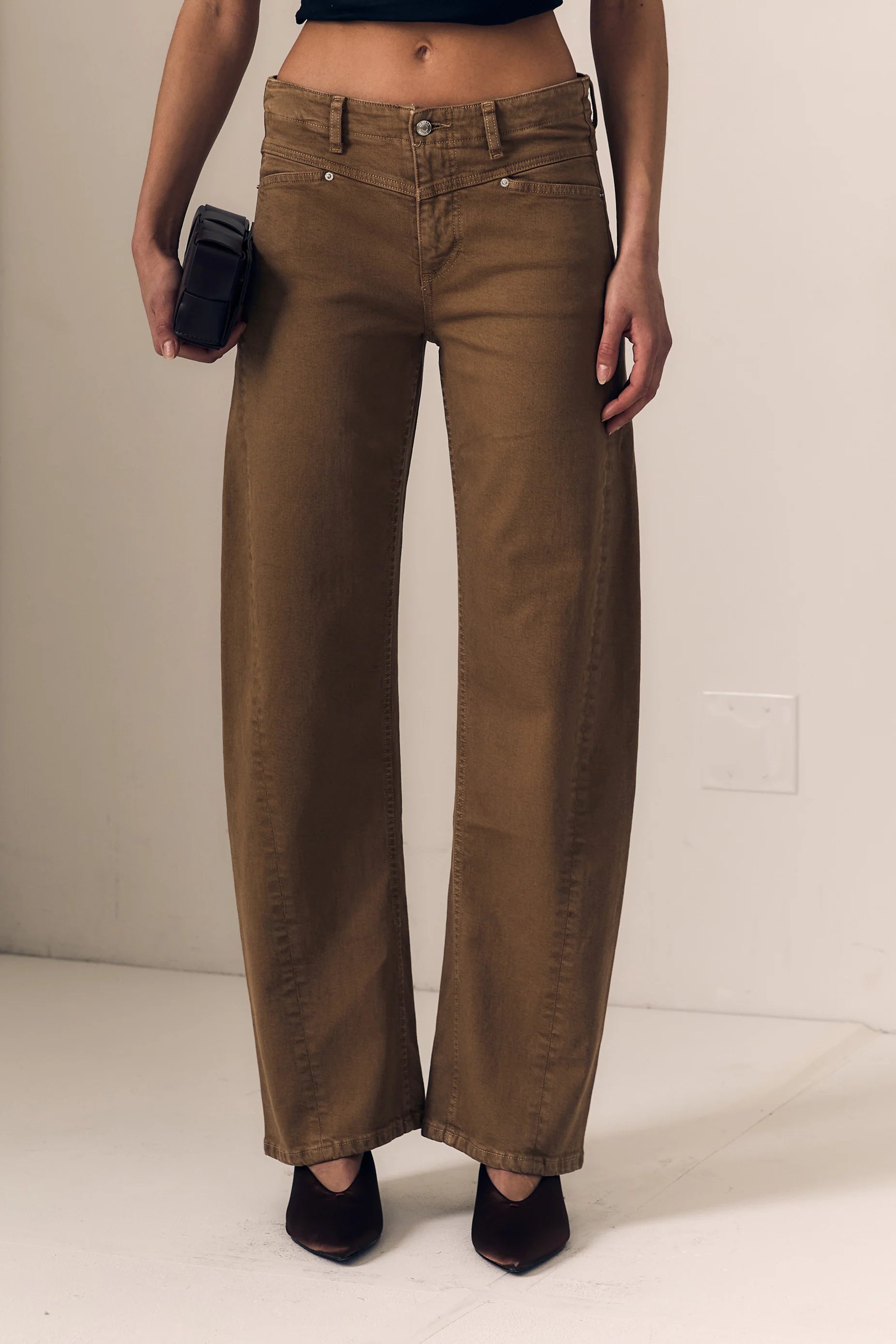A person wearing brown wide-leg cropped jeans with a black crop top and black pointed shoes, holding a black phone against a plain off-white wall.
