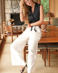 Woman in black top and white pants sitting in a cozy room with stone walls and wooden furniture.