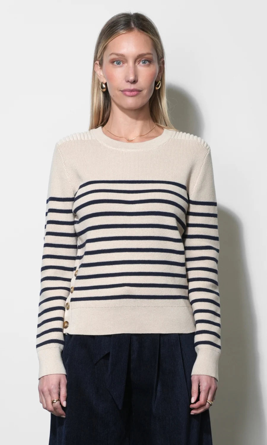 A woman modeling a cream-colored knit sweater with navy blue horizontal stripes across the chest and sleeves, paired with dark navy trousers, against a plain off-white background.