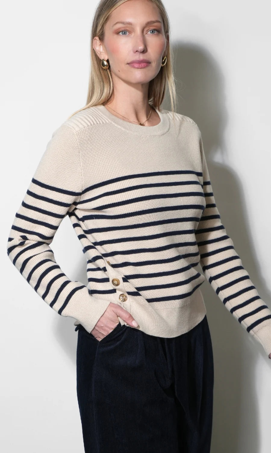 A woman wearing a cream and navy striped knit sweater with a buttoned asymmetrical hem, paired with dark trousers, standing against a plain light gray wall.