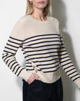 A woman wearing a cream and navy striped knit sweater with a buttoned asymmetrical hem, paired with dark trousers, standing against a plain light gray wall.