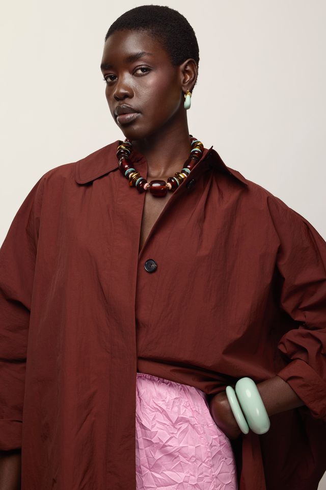 A model wearing an oversized rust-brown button-up shirt and a pink textured skirt, accessorized with a Carmine Collar Necklace and light green bangles, against a plain beige background.