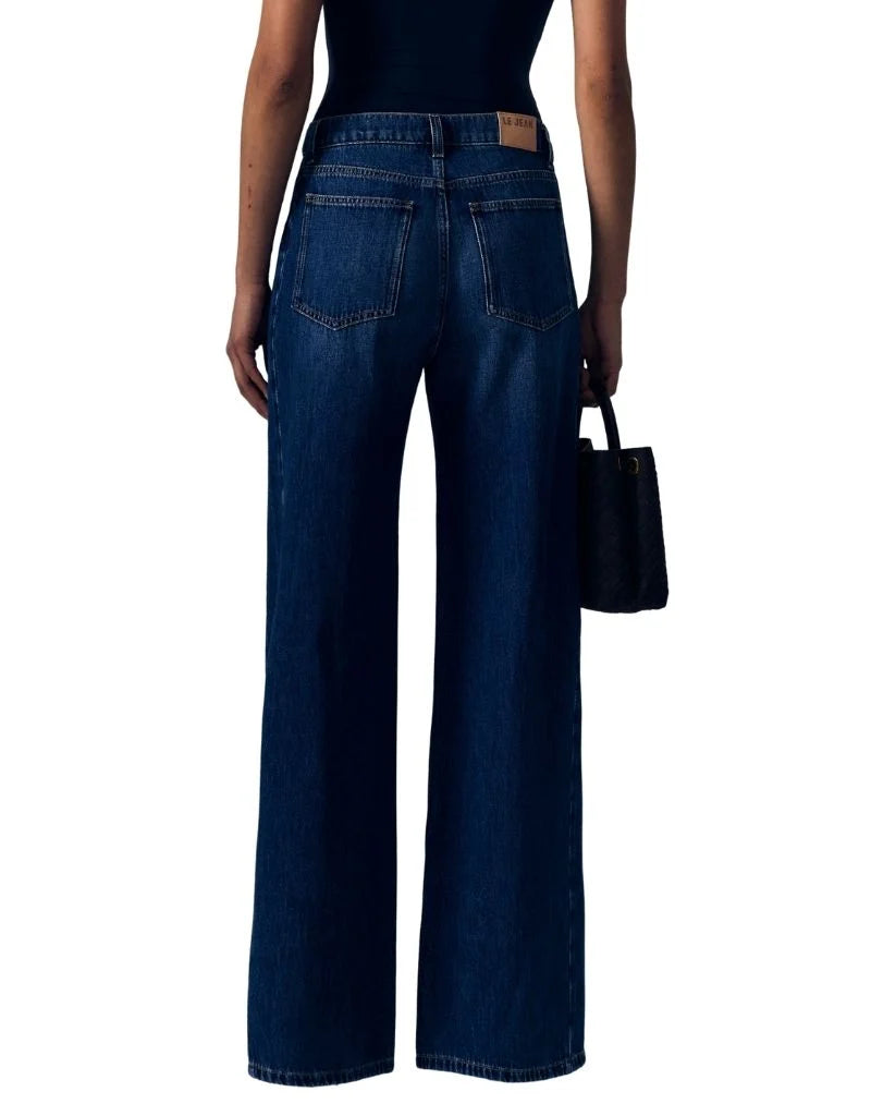 Rear view of a model wearing dark blue wide-leg jeans with a black top and holding a black handbag against a white background.