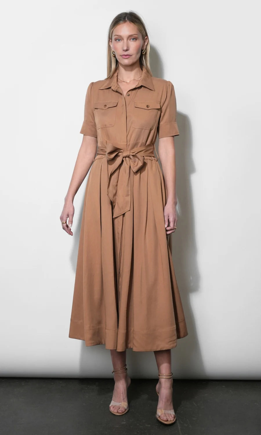 A woman modeling a tan short-sleeve midi dress with a button-up collar, chest flap pockets, and a matching fabric tie belt at the waist, standing against a plain light gray wall.