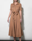 A woman modeling a tan short-sleeve midi dress with a button-up collar, chest flap pockets, and a matching fabric tie belt at the waist, standing against a plain light gray wall.