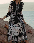 Person wearing a black and white patterned dress sitting on a rock by the water.