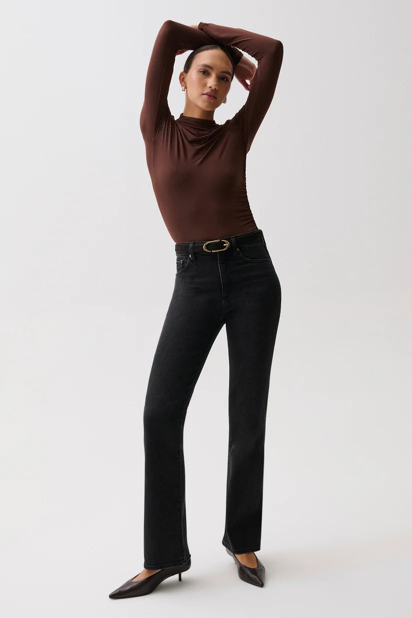 A model posing in a studio setting, wearing a dark brown mock-neck long-sleeve top, black high-waisted flared jeans with a gold-tone belt, and black pointed-toe heels.