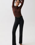A model posing in a studio setting, wearing a dark brown mock-neck long-sleeve top, black high-waisted flared jeans with a gold-tone belt, and black pointed-toe heels.