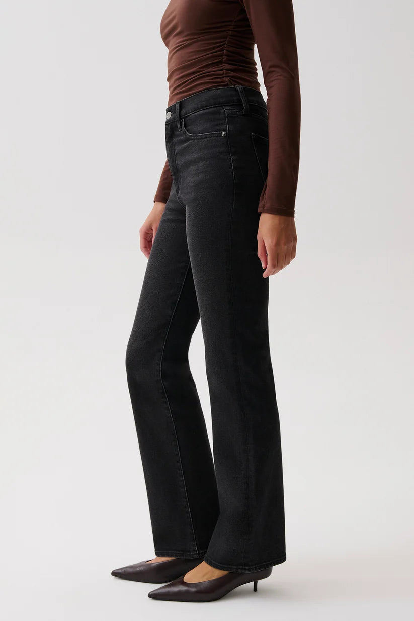 A model wearing black wide-leg flare jeans, a brown long-sleeve top, and dark brown pointed-toe flats, shown from the waist down against a light gray studio background.