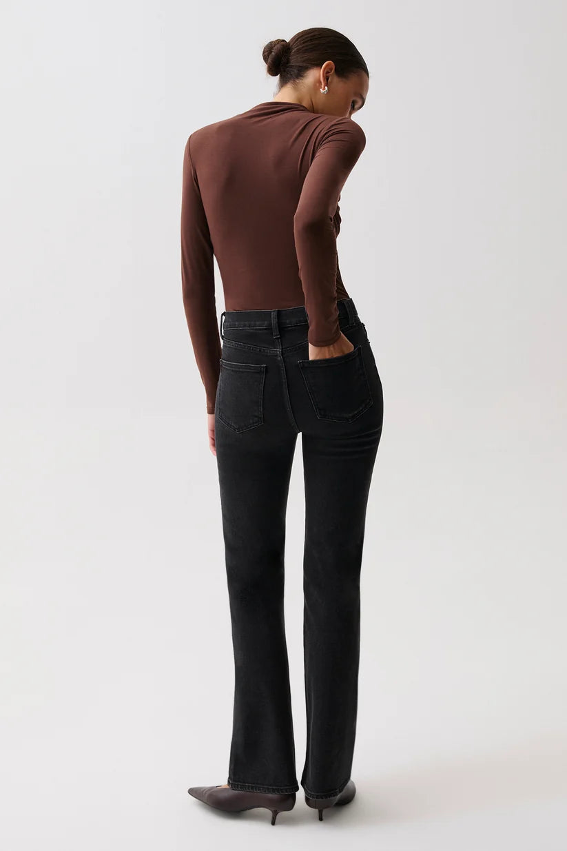 A model shown from the back wearing a brown long-sleeve top, black flare jeans, and dark pointed-toe heels against a plain light gray studio background.