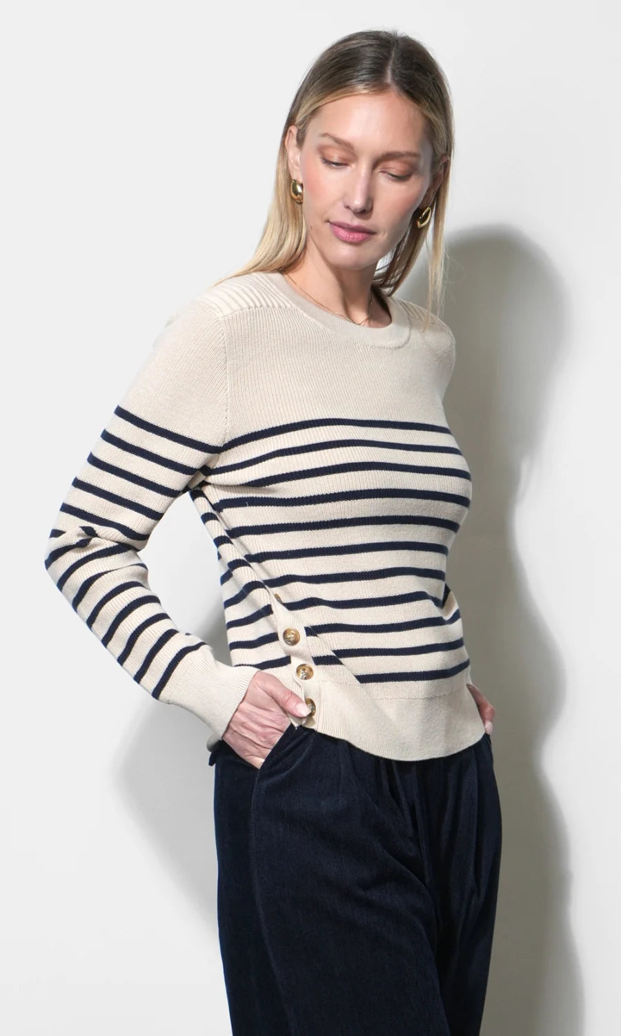A woman wearing a cream and navy striped knit sweater with gold buttons on the front, paired with dark navy pants, standing against a light gray wall.