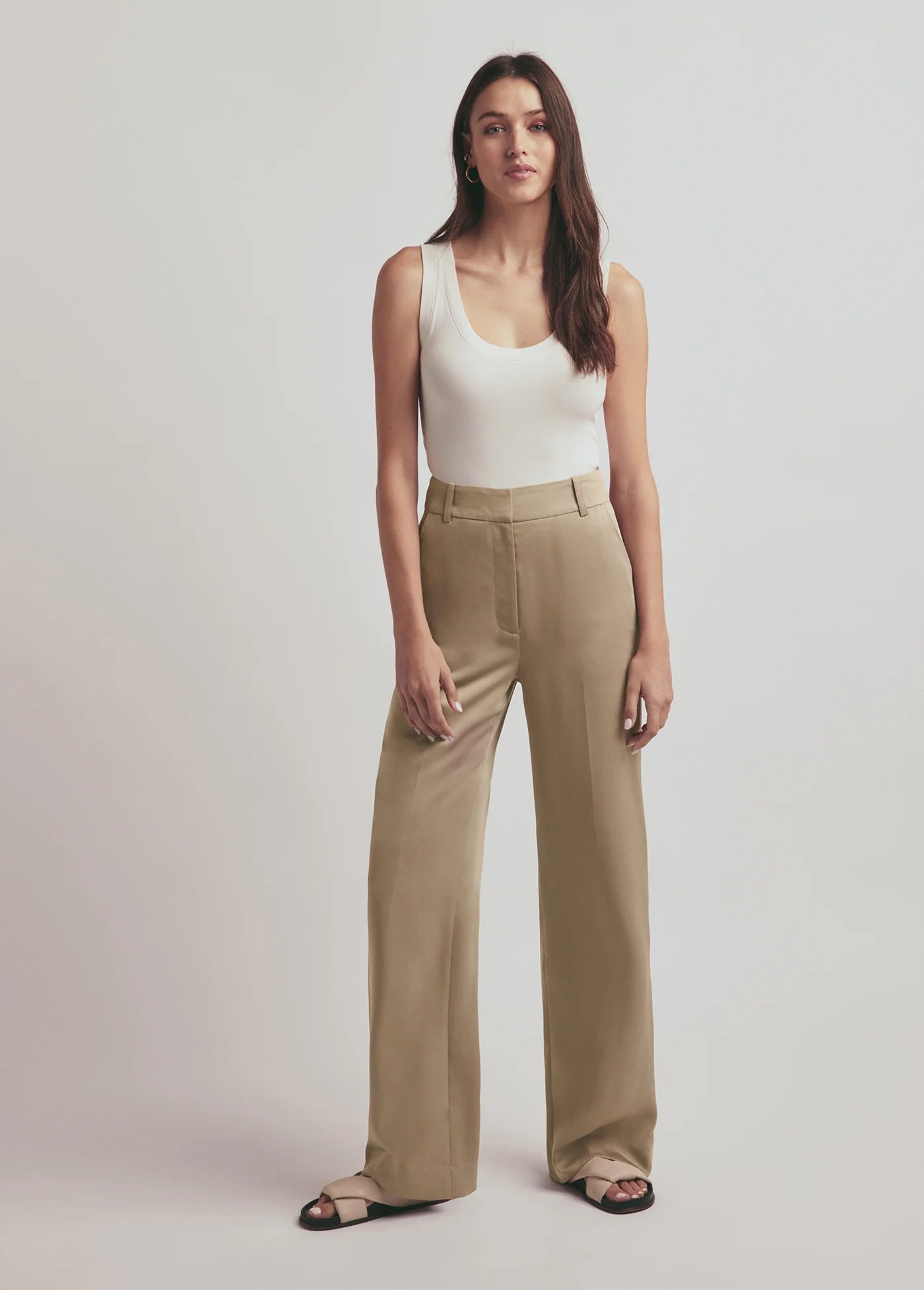 Woman wearing a white tank top and beige pants on a plain background, front view