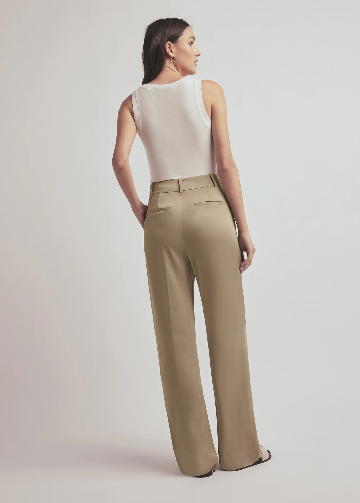 Woman wearing a white sleeveless top and beige high-waisted pants on a plain background, back view