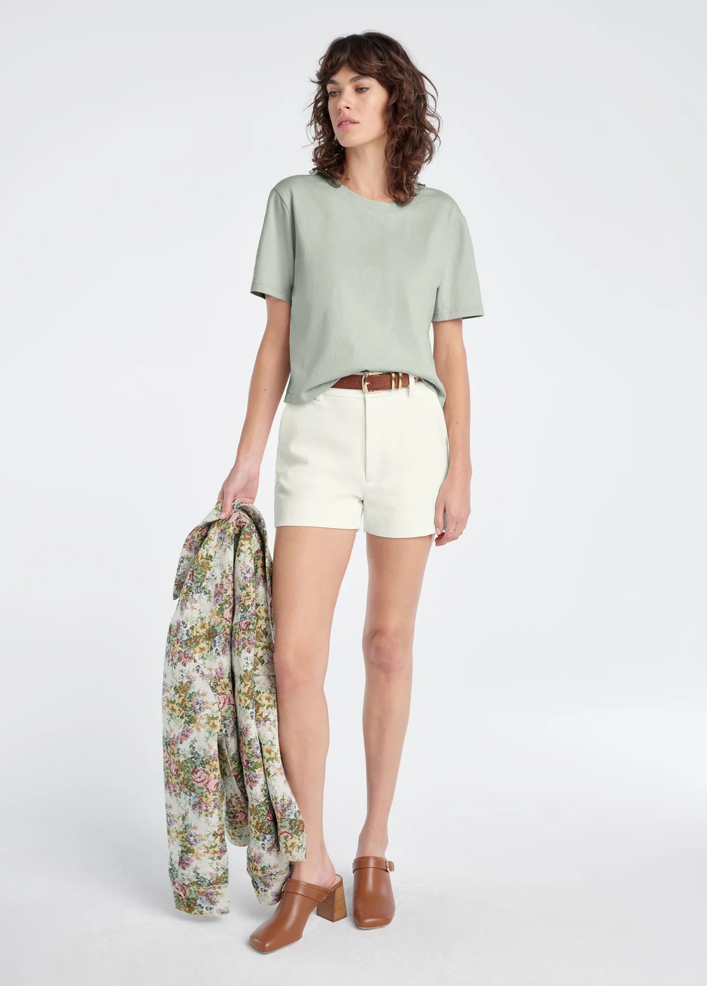 Woman holding a floral jacket, wearing a light green top and white shorts on a white background