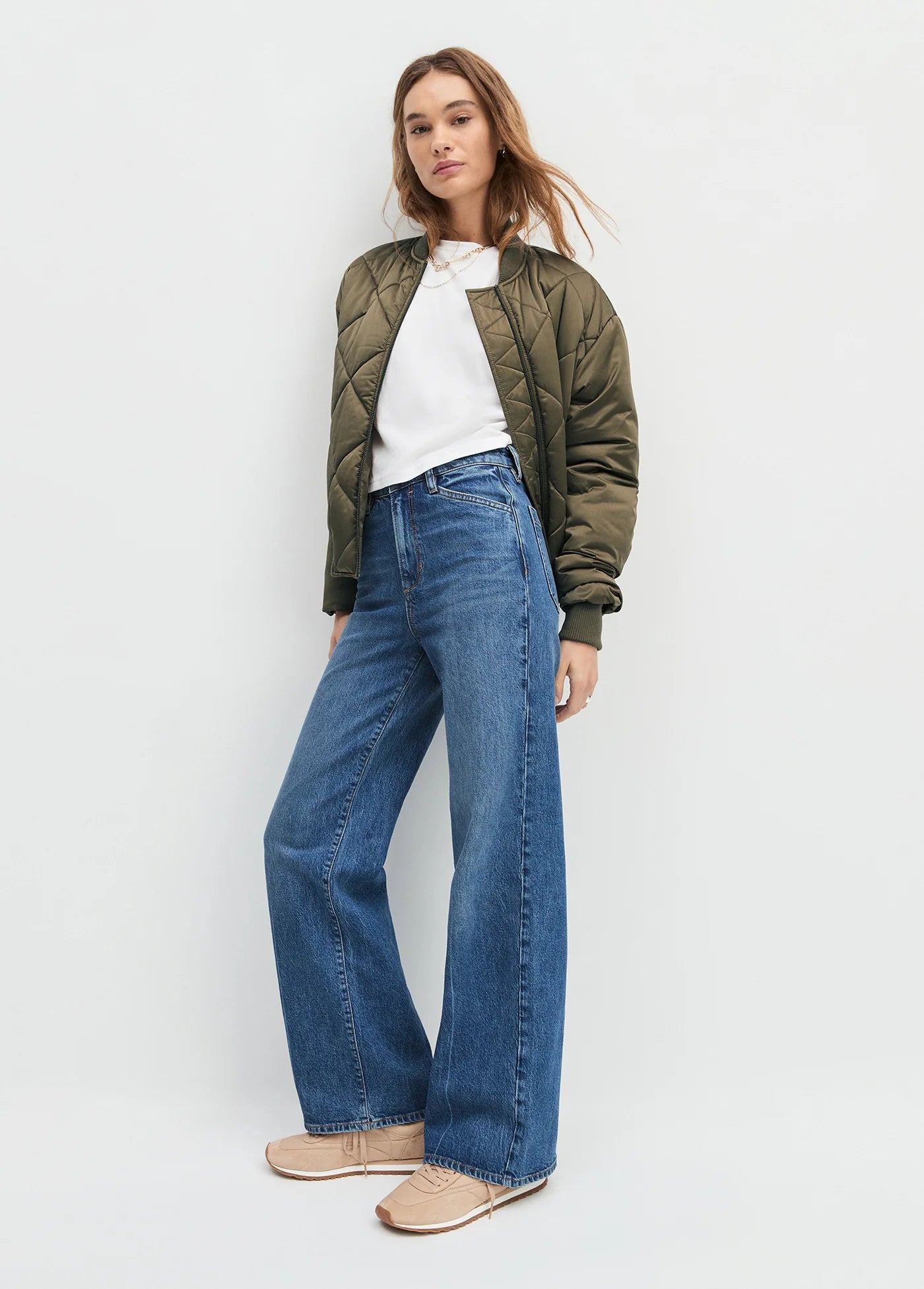 Woman wearing a green bomber jacket, white shirt, blue jeans, and beige shoes on a light gray background, side view