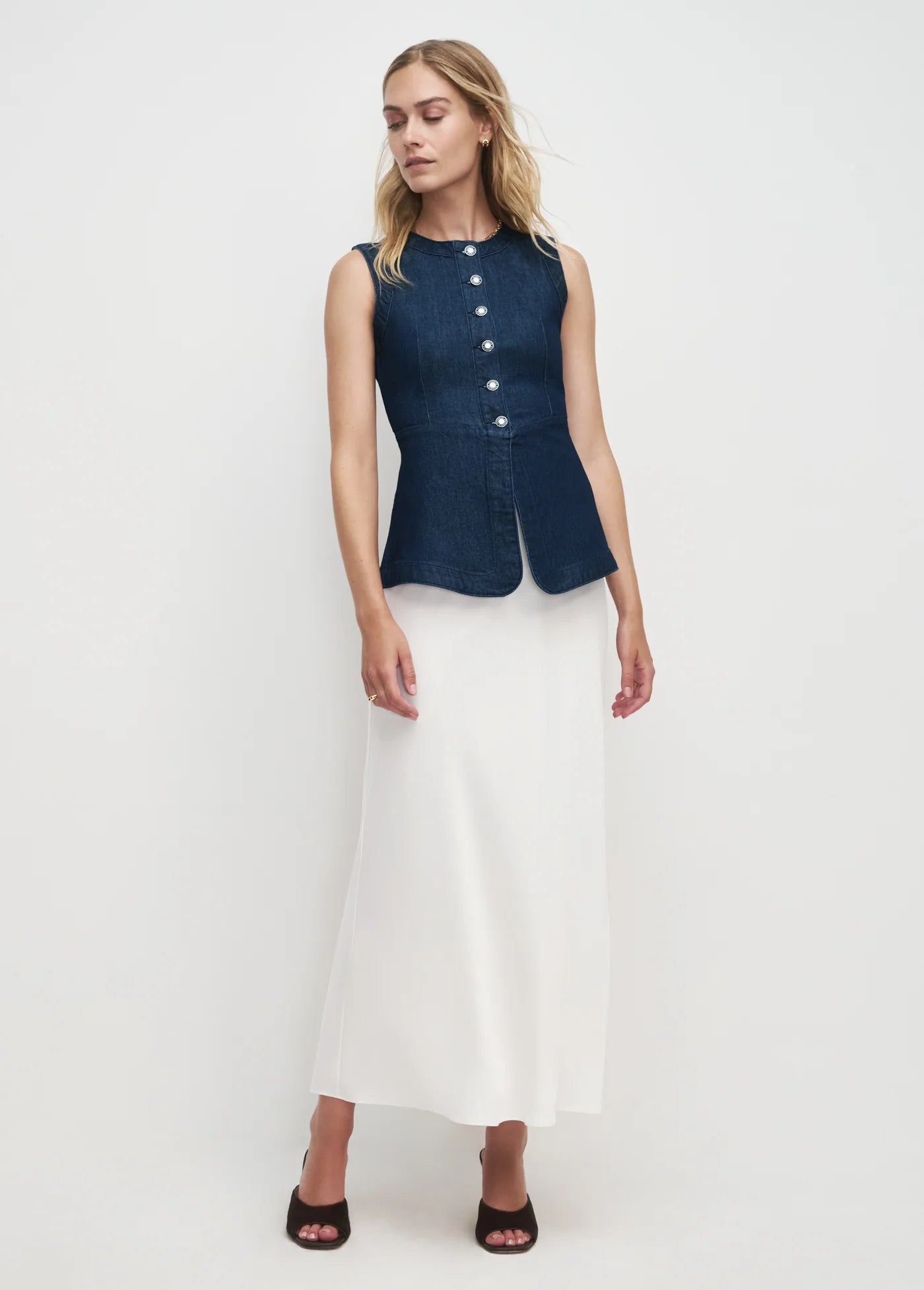 Woman wearing a navy sleeveless button-up top and white pants on a white background