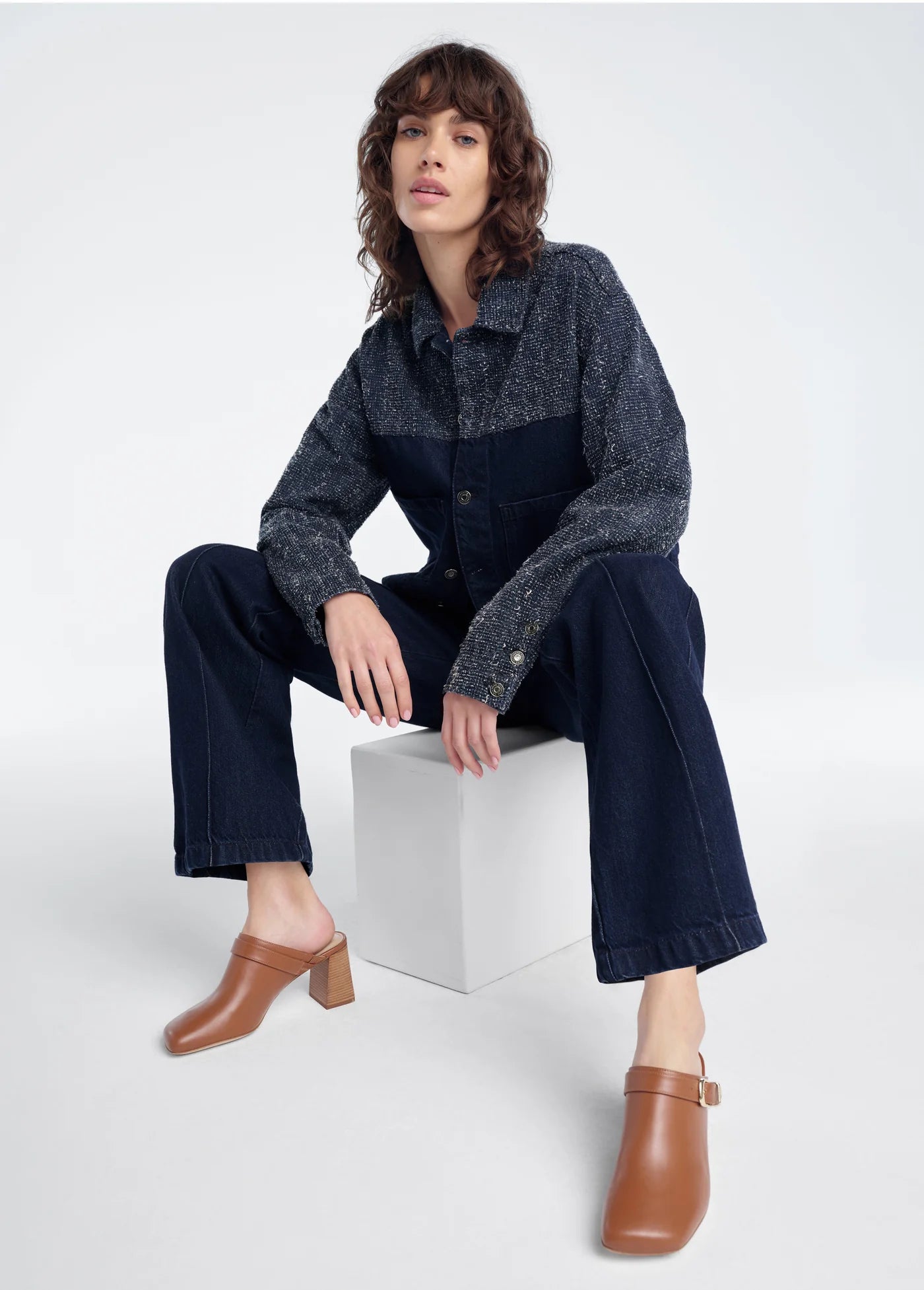 Woman sitting on a white cube wearing a dark denim outfit with a patterned shirt.