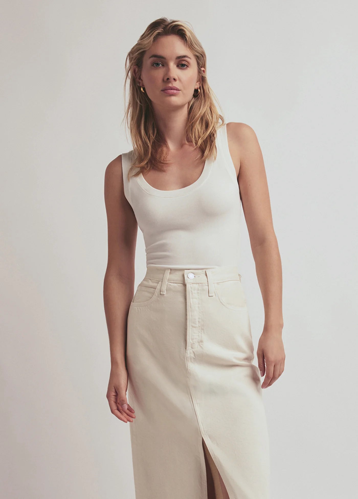 Woman wearing a white tank top and beige skirt against a plain background