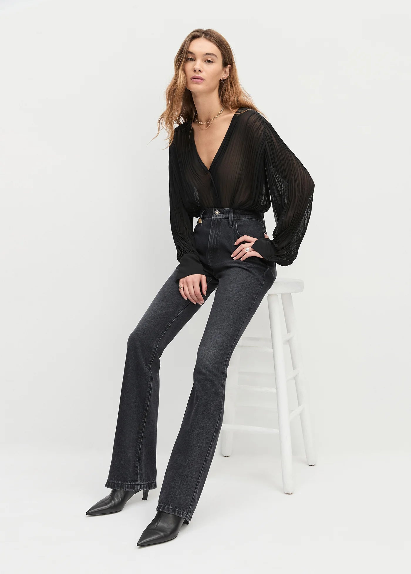 Woman wearing a black blouse and dark jeans sitting on a white stool against a white background