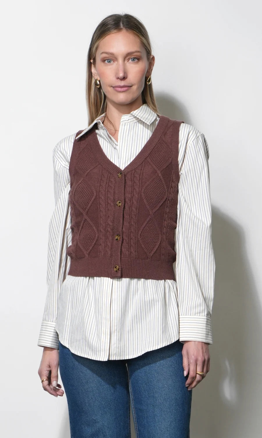A woman modeling a brown cable-knit vest over a white and navy striped button-down shirt and blue jeans, standing against a plain light gray wall.