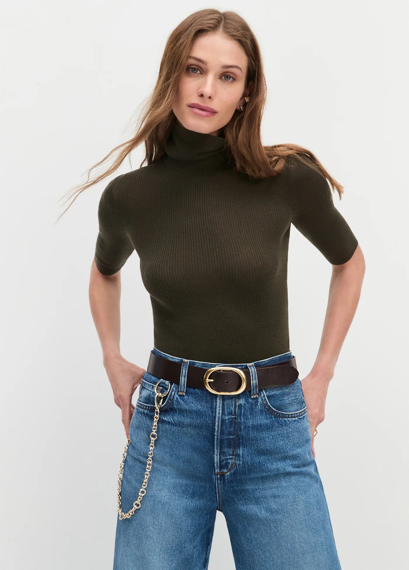 Woman wearing a dark green turtleneck and blue jeans with a belt on a light gray background