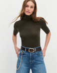 Woman wearing a dark green turtleneck and blue jeans with a belt on a light gray background
