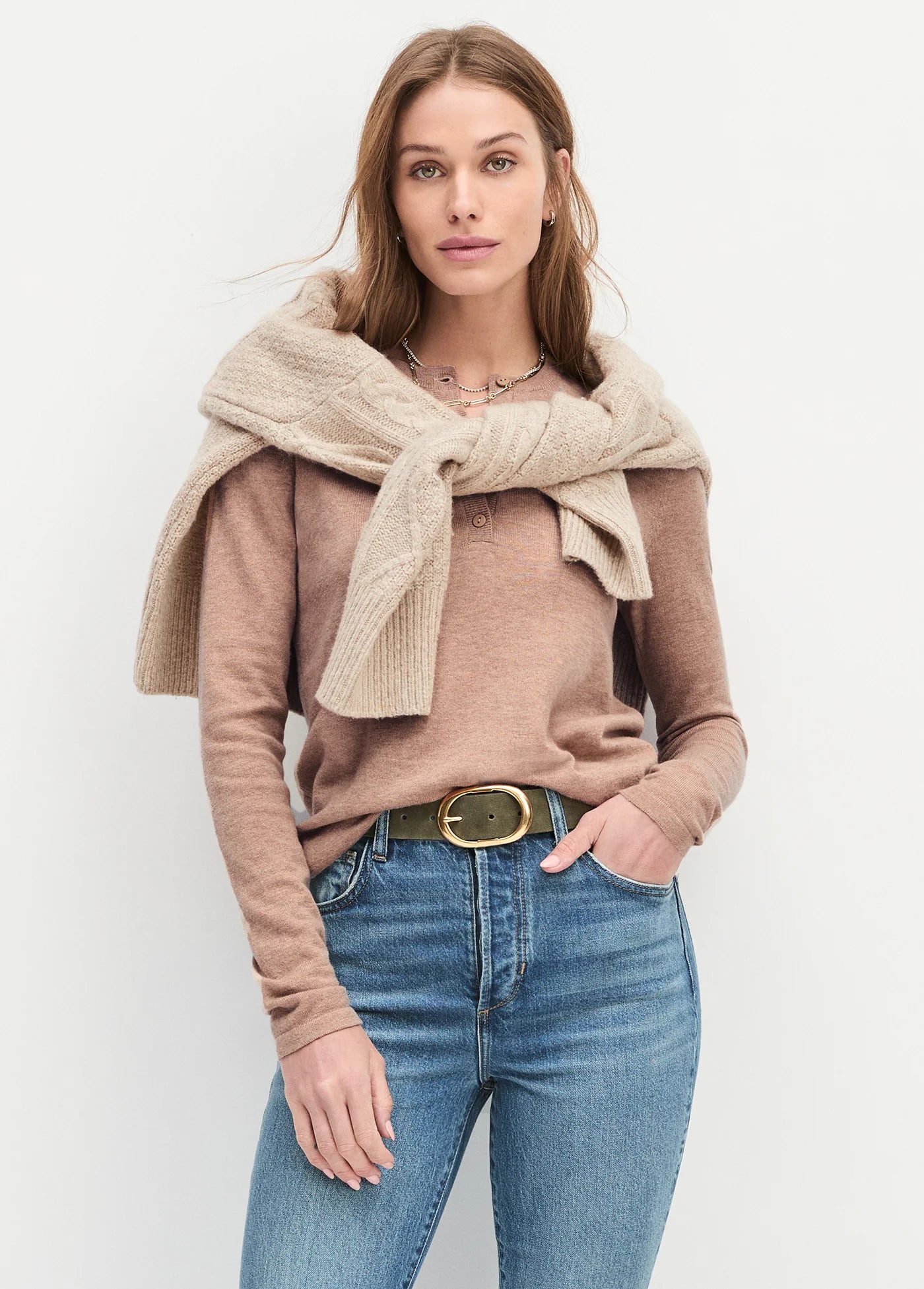 Woman wearing a beige sweater, scarf, and blue jeans against a white background