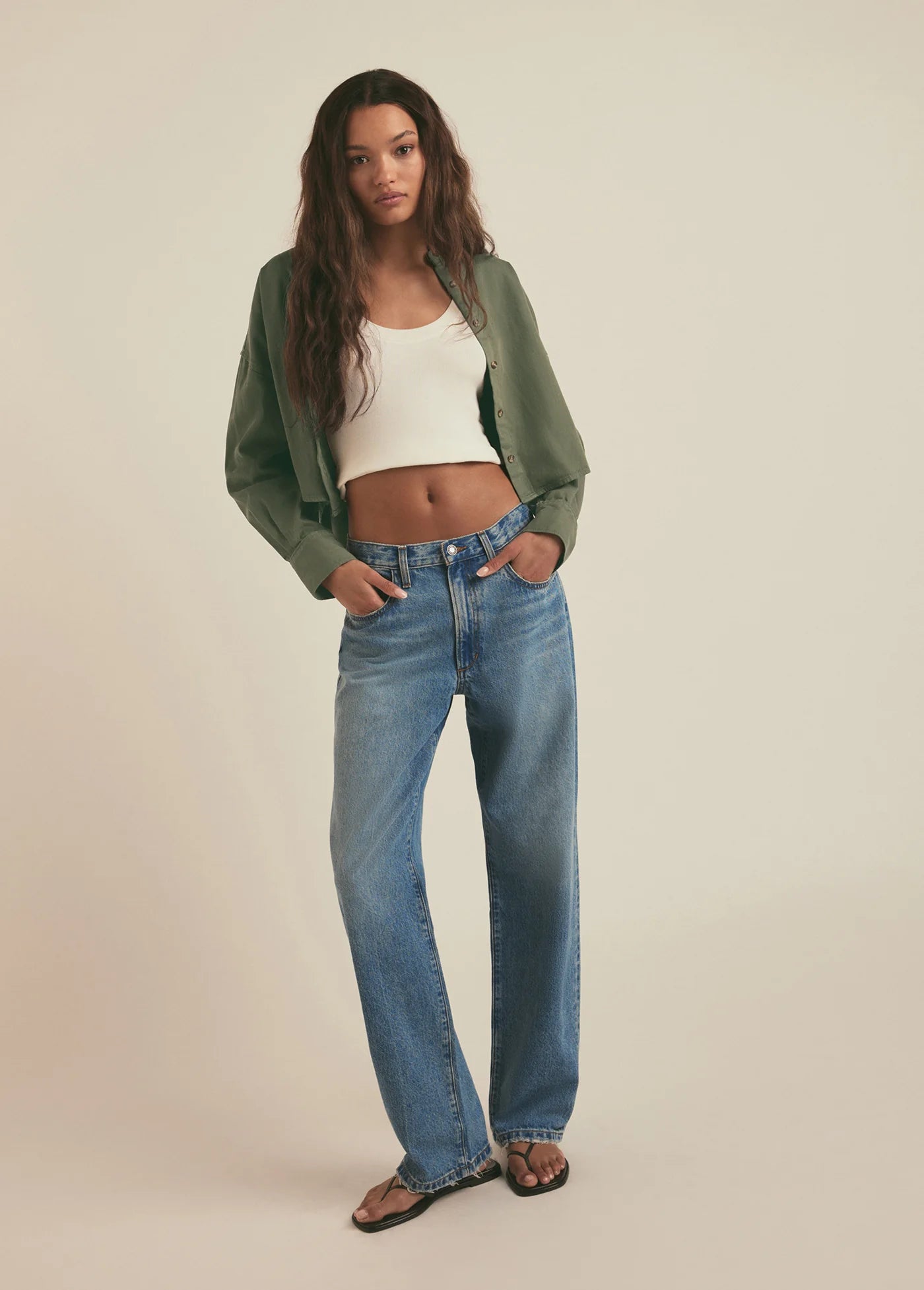 Woman wearing a green jacket, white top, and blue jeans on a beige background