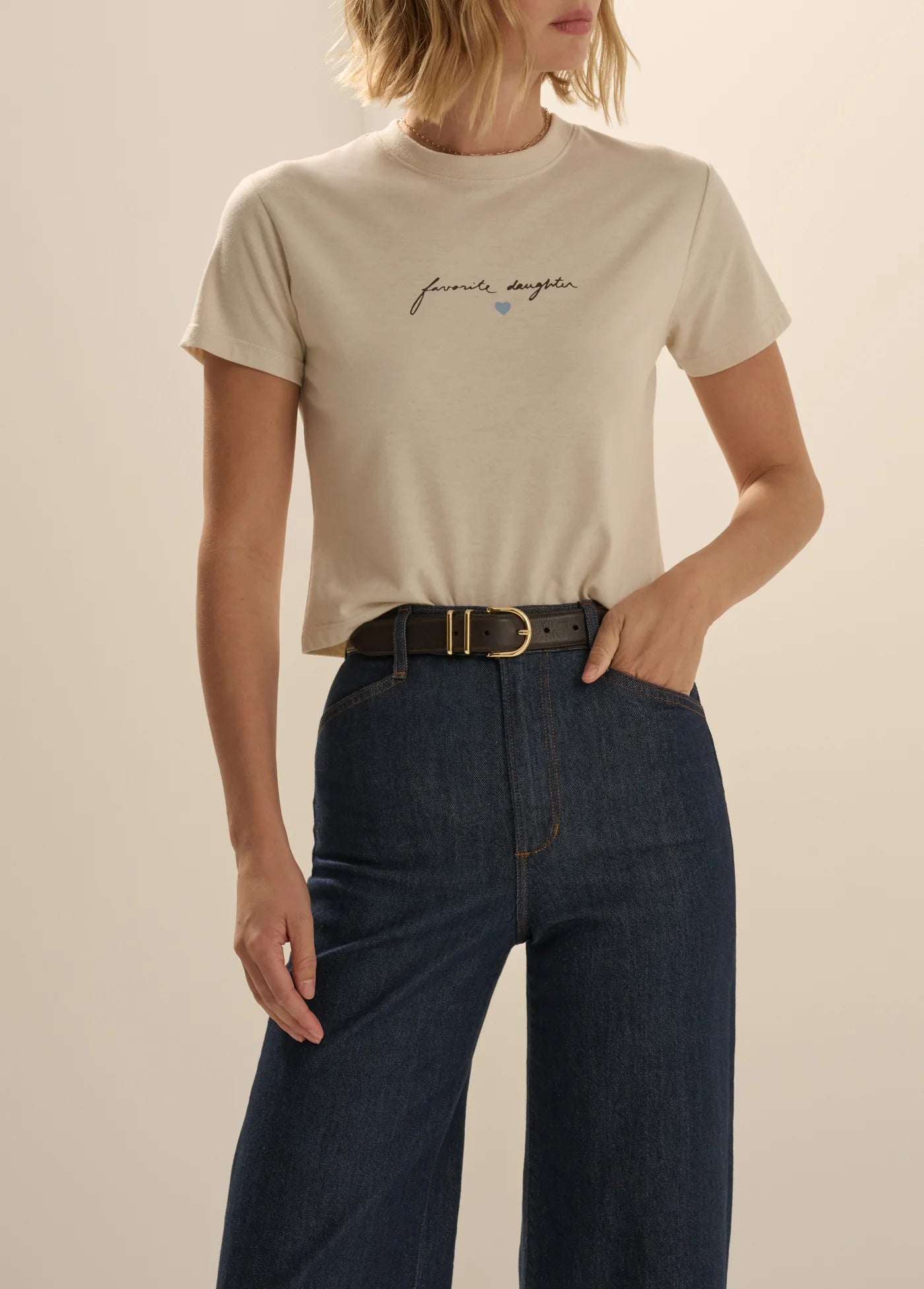 A woman wearing a light beige short-sleeve t-shirt with a small black script graphic and a tiny blue heart on the chest, paired with dark blue jeans and a black belt, against a solid beige background.