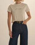 A woman wearing a light beige short-sleeve t-shirt with a small black script graphic and a tiny blue heart on the chest, paired with dark blue jeans and a black belt, against a solid beige background.