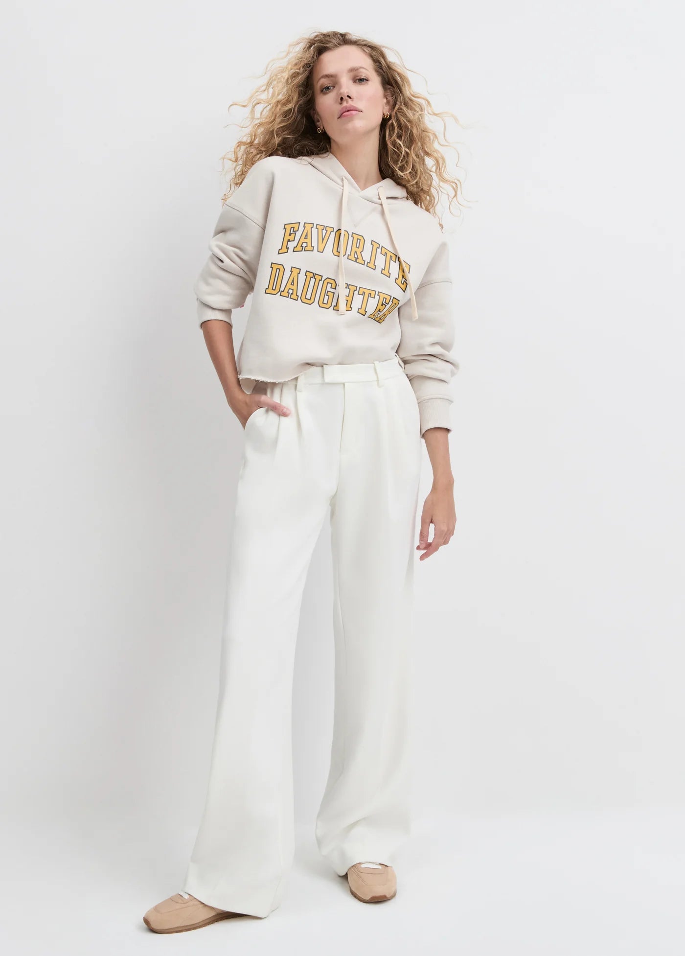Woman wearing a beige hoodie with 'Favorite Daughter' text and white pants on a light gray background