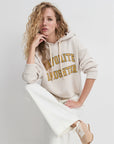 Woman wearing a beige hoodie with 'FAVORITE DAUGHTER' text, white pants, and beige shoes on a light gray background