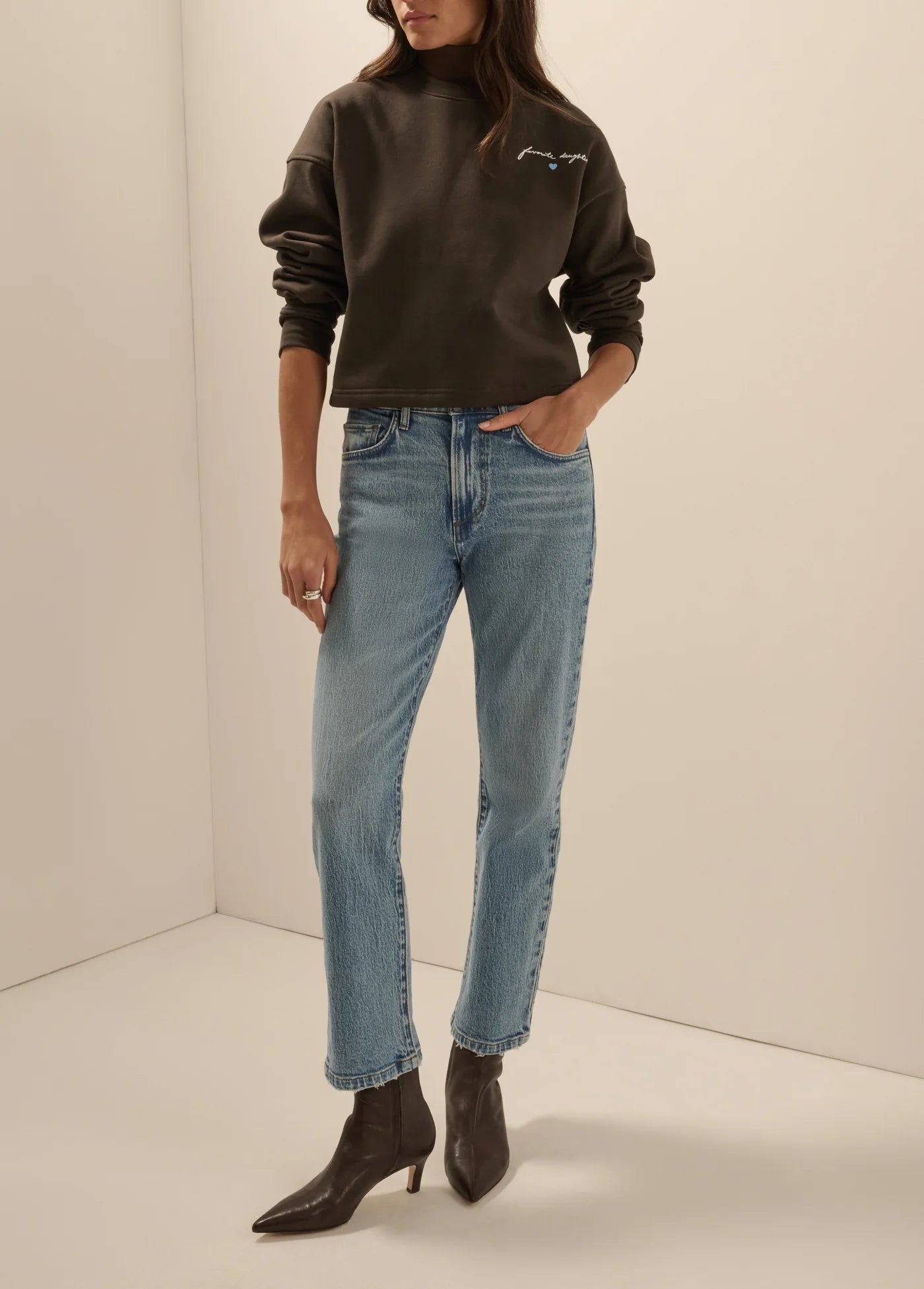 A model wearing a dark brown sweatshirt with a small embroidered logo on the chest, paired with light-wash blue jeans and dark brown pointed-toe ankle boots, standing in a minimalist cream-colored studio corner.