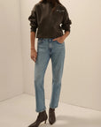 A model wearing a dark brown sweatshirt with a small embroidered logo on the chest, paired with light-wash blue jeans and dark brown pointed-toe ankle boots, standing in a minimalist cream-colored studio corner.