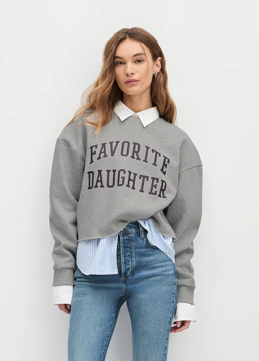 Woman wearing a gray sweatshirt with 'Favorite Daughter' text, light blue shirt, and jeans on a white background front view