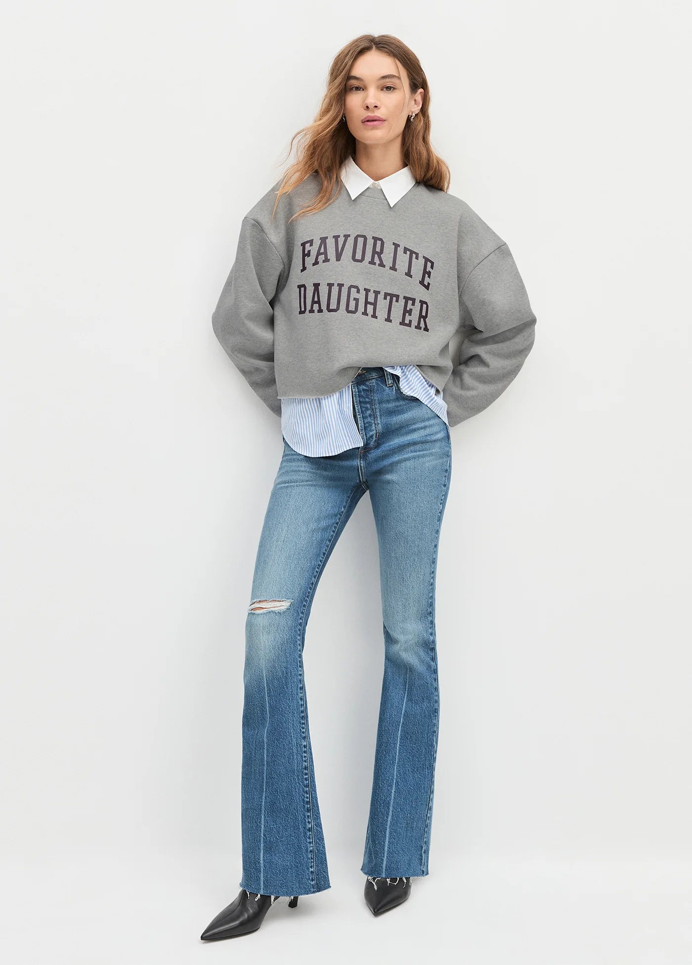 Woman wearing a gray 'Favorite Daughter' sweatshirt with a white shirt and blue jeans on a light gray background