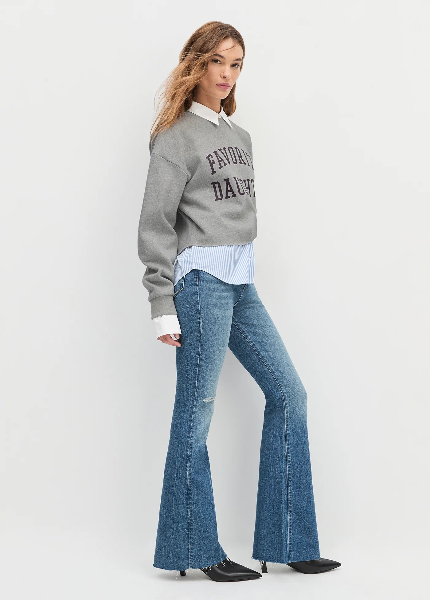 Woman wearing a gray sweatshirt with text, white shirt, and blue flared jeans on a white background side view