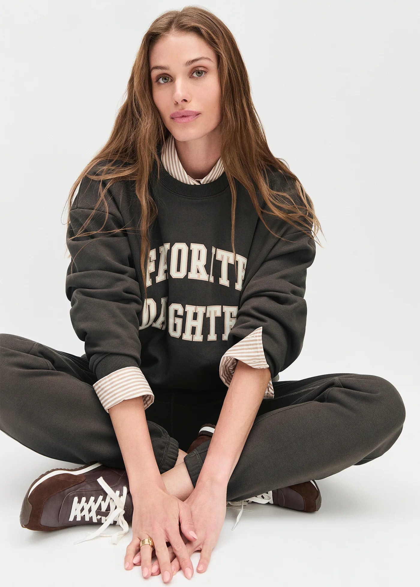 model wearing collegiate sweatshirt with brown pants and brown shoes sitting on the ground with a white background