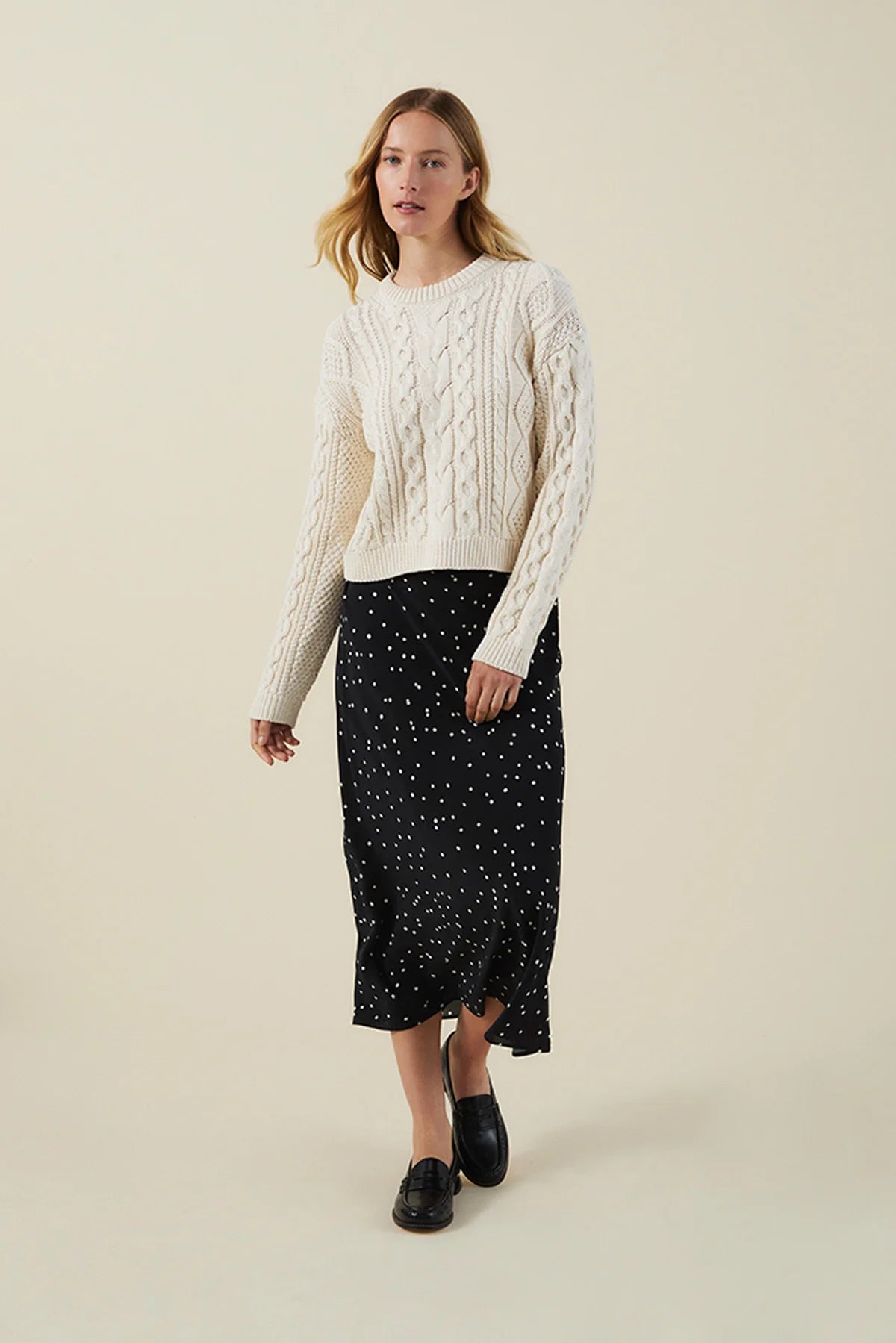 A woman modeling a cream cable-knit sweater and a black midi skirt with small white polka dots, paired with black loafers, against a solid light beige background.