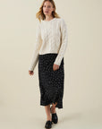A woman modeling a cream cable-knit sweater and a black midi skirt with small white polka dots, paired with black loafers, against a solid light beige background.
