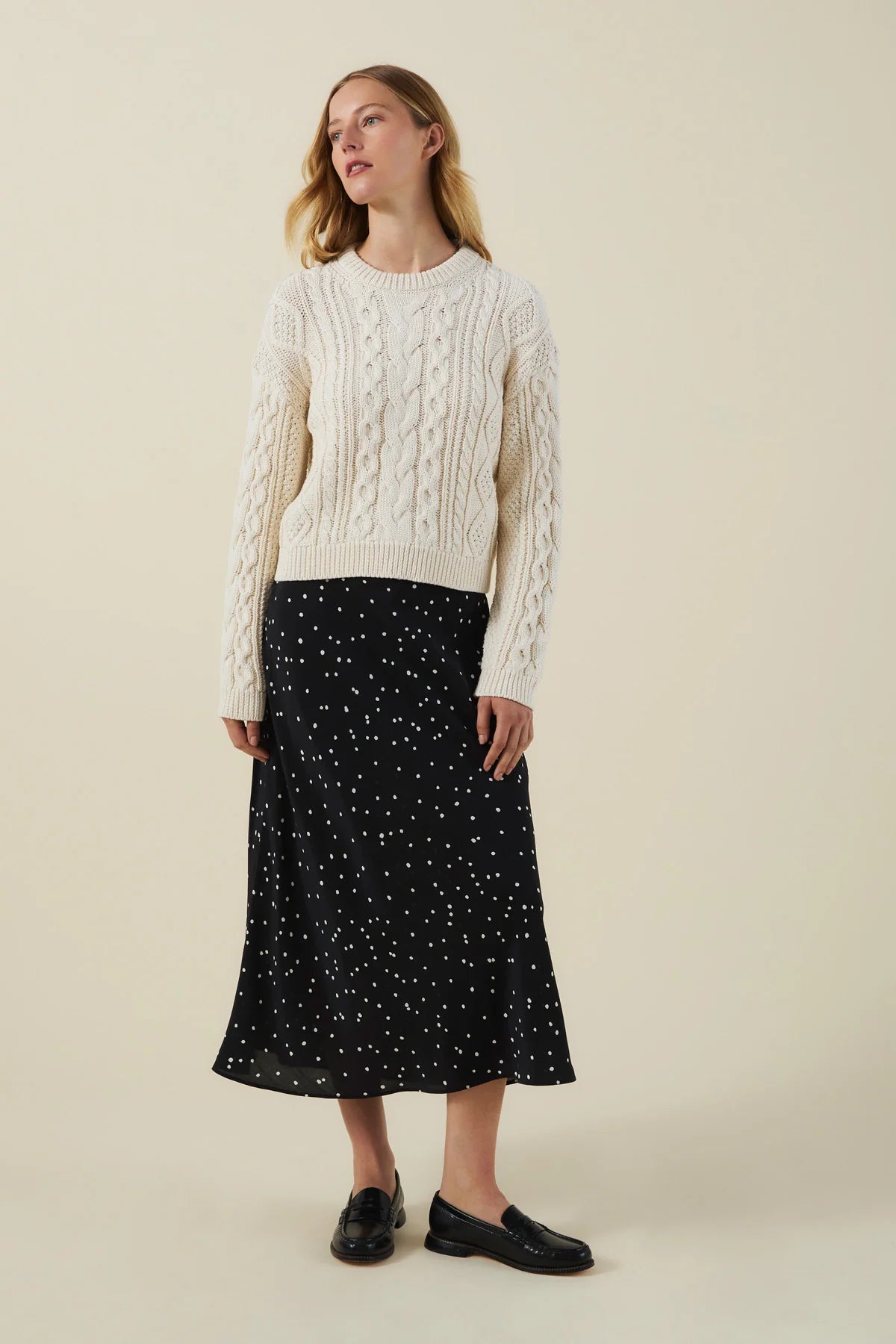A woman stands against a plain beige studio background wearing a cream cable-knit sweater, a black midi skirt with small white polka dots, and black loafers.