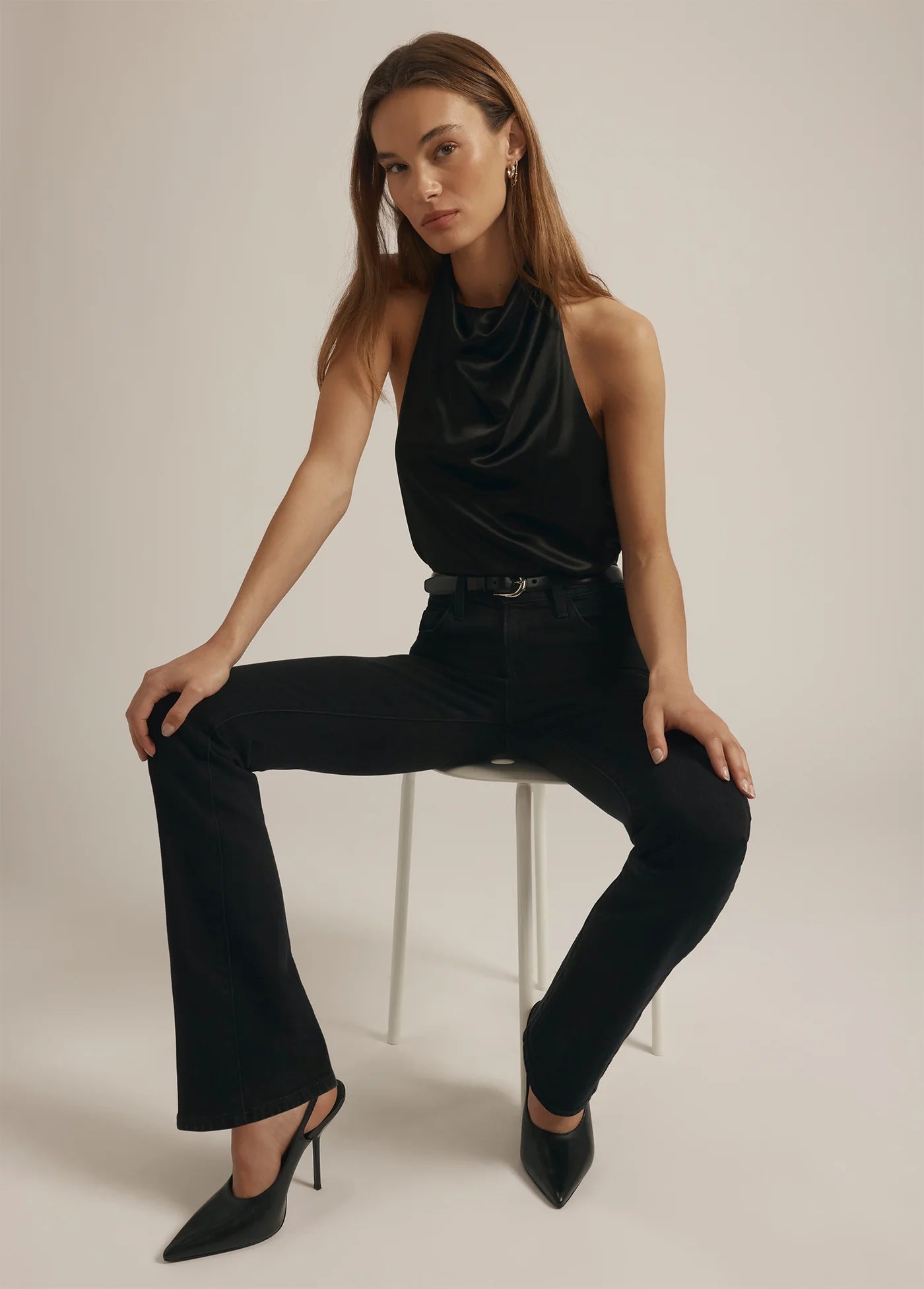 Woman sitting on a stool wearing a black sleeveless top and black pants against a plain background