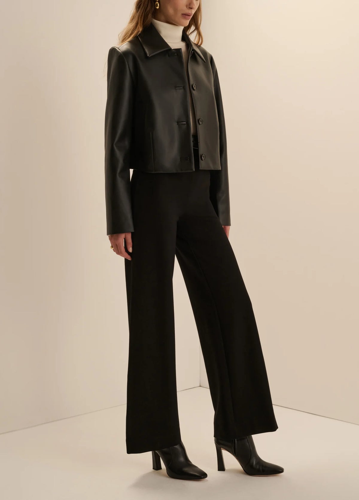 A model wearing a black cropped faux-leather jacket with structured shoulders, black wide-leg trousers, a white mock-neck top, and black pointed-toe ankle boots, standing against a warm off-white studio background.