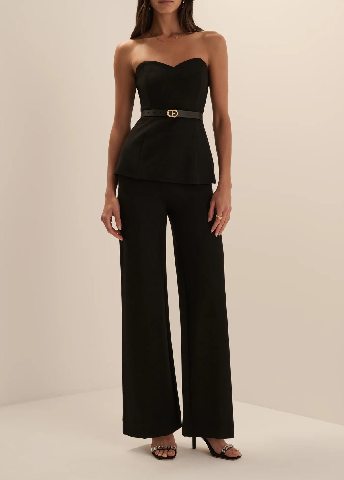 A woman modeling a black strapless top and matching wide-leg trousers set, paired with black heeled sandals, in a minimalist cream studio setting.