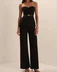 A woman modeling a black strapless top and matching wide-leg trousers set, paired with black heeled sandals, in a minimalist cream studio setting.