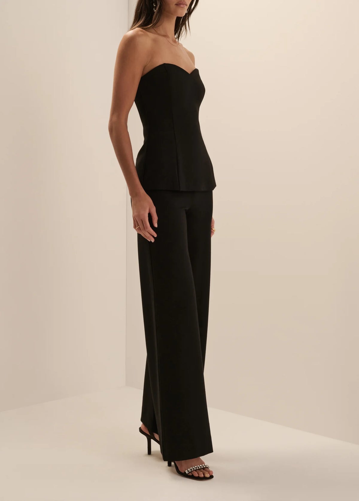 A woman modeling a black strapless top and matching wide-leg trousers set, paired with black heeled sandals, in a minimalist cream studio setting.